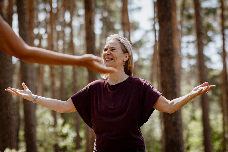 Eine lächelnde Frau mit ergrauenden Haaren und dunkelrotem Oberteil steht mit weit geöffneten, ausgebreiteten Armen im sonnigen Kiefernwald. Im linken Vordergrund ragt unscharf der Arm einer weiteren Person ins Bild.