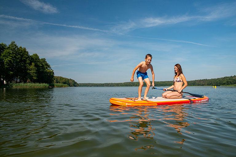 Bei blauem Himmel befinden sich eine Frau und ein Junge auf einem leuchtend orange-weißen Stand-Up-Paddleboard mitten auf einem See. Die Frau kniet im hinteren Teil des Boards und hält quer ein Stechpaddel in den Händen, während der Junge im vorderen Teil steht und die Balance hält. Im Hintergrund sind grüne, von Bäumen gesäumte Ufer zu sehen.
