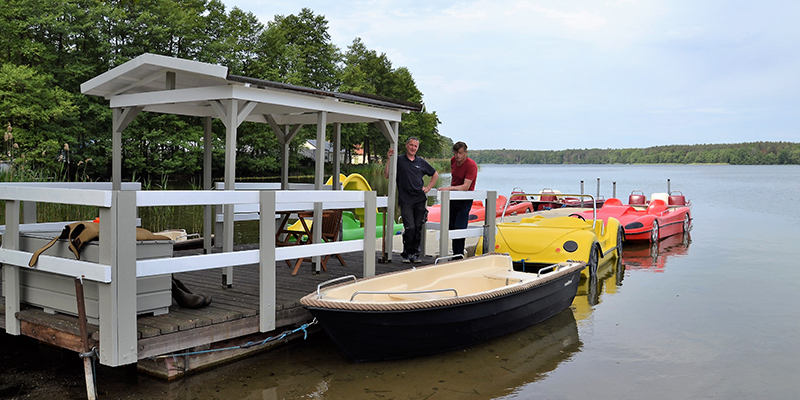 Zwei Männer stehen auf einem Holzsteg mit einem kleinen, überdachten Unterstand an einem ruhigen See. Neben dem Steg liegen mehrere kleine Wasserfahrzeuge: Im Vordergrund schwimmt ein dunkles Ruderboot mit heller Innenschale, während dahinter auffällige gelbe und rote Tretboote – teilweise im Auto-Design – festgemacht sind. Das gegenüberliegende Ufer im Hintergrund ist von einem dichten grünen Wald gesäumt.