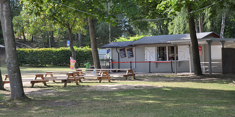 Ein hellgrauer Imbiss-Pavillon mit geöffnetem Verkaufsfenster und zwei dunklen Menütafeln an der Fassade steht unter großen, schattenspendenden Bäumen. Auf der Rasenfläche im Vordergrund sind mehrere hölzerne Picknicktische mit dazugehörigen Bänken aufgestellt. Eine rote Absperrung führt als Wegweiser zum Schalter, während im linken Hintergrund eine dichte grüne Hecke zu erkennen ist.