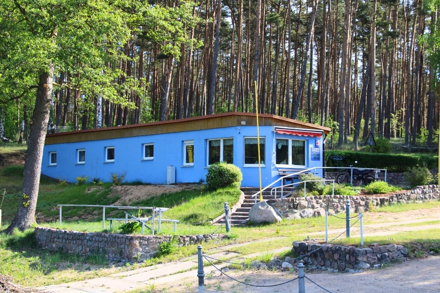 Ein langgestrecktes, hellblau gestrichenes Gebäude mit mehreren weißen Fenstern steht auf einer leichten Anhöhe vor einem dichten Kiefernwald. An der rechten Gebäudeseite ist ein Schild mit einem roten Kreuz angebracht, und über einem der Fenster ist eine rote Markise gespannt. Eine kurze Steintreppe führt den grasbewachsenen Hang hinauf zum Gebäude, während im Vordergrund flache Mauern aus Natursteinen das Gelände strukturieren.