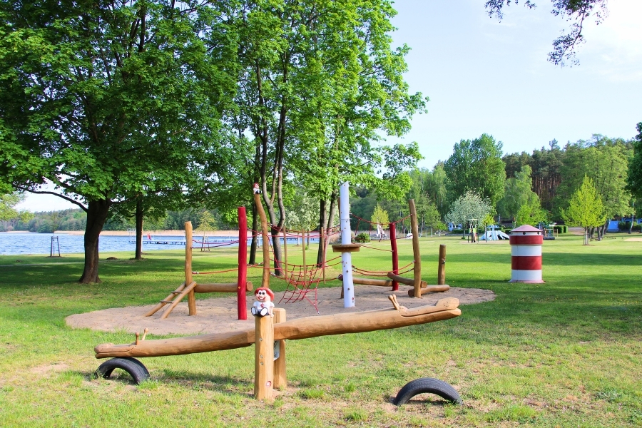 Ein weitläufiger Spielplatz auf einer grünen Rasenfläche mit großen, schattenspendenden Bäumen in der Nähe eines Sees. Im Vordergrund steht eine massive Holzwippe, die an den Enden durch halb eingegrabene Autoreifen abgefedert wird; auf dem Mittelpfosten sitzt eine kleine Deko-Figur. Dahinter befindet sich auf einer Sandfläche ein Klettergerüst aus naturbelassenen Holzpfosten und roten Seilen. Rechts daneben steht eine kleine, rot-weiß gestreifte Säule, die optisch an einen Leuchtturm erinnert.