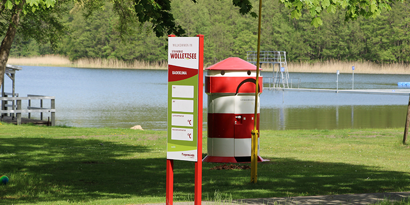 Eine rund geformte Umkleidekabine, die optisch wie ein rot-weiß gestreifter Leuchtturm gestaltet ist, steht auf einer Wiese im Strandbad Wolletzsee. Davor befindet sich eine aufrecht stehende, rot-weiße Informationstafel zum Badeklima mit dem Logo des Strandbads. Im Hintergrund erstrecken sich das Ufer des Sees, ein Steg sowie ein dichter Wald unter einem klaren Himmel.