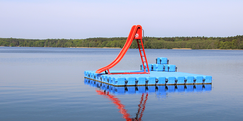 Eine rote Wasserrutsche steht auf einer blauen, schwimmenden Plattform aus Kunststoffelementen, die mitten auf dem ruhigen Wolletzsee im Strandbad Wolletzsee treibt. Im Hintergrund ist das flache, bewaldete Ufer unter einem hellen Himmel zu sehen.