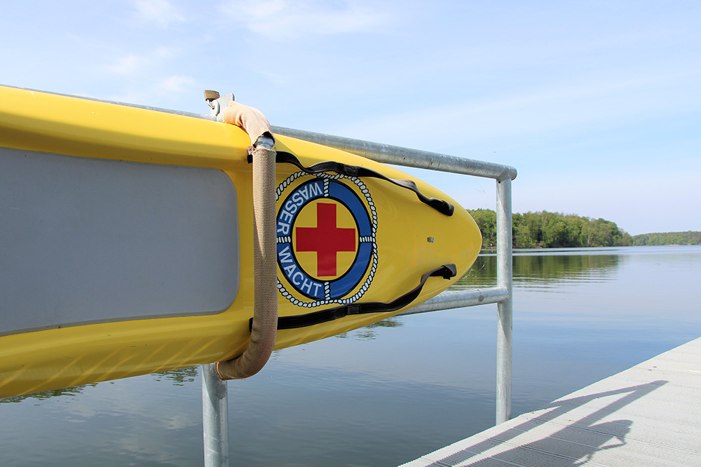 Ein gelbes Rettungsboard der Wasserwacht mit einem blauen Logo und rotem Kreuz ist an einer metallenen Steganlage im Strandbad Wolletzsee befestigt. Im Hintergrund sind das ruhige Wasser des Sees und das bewaldete Ufer zu sehen.