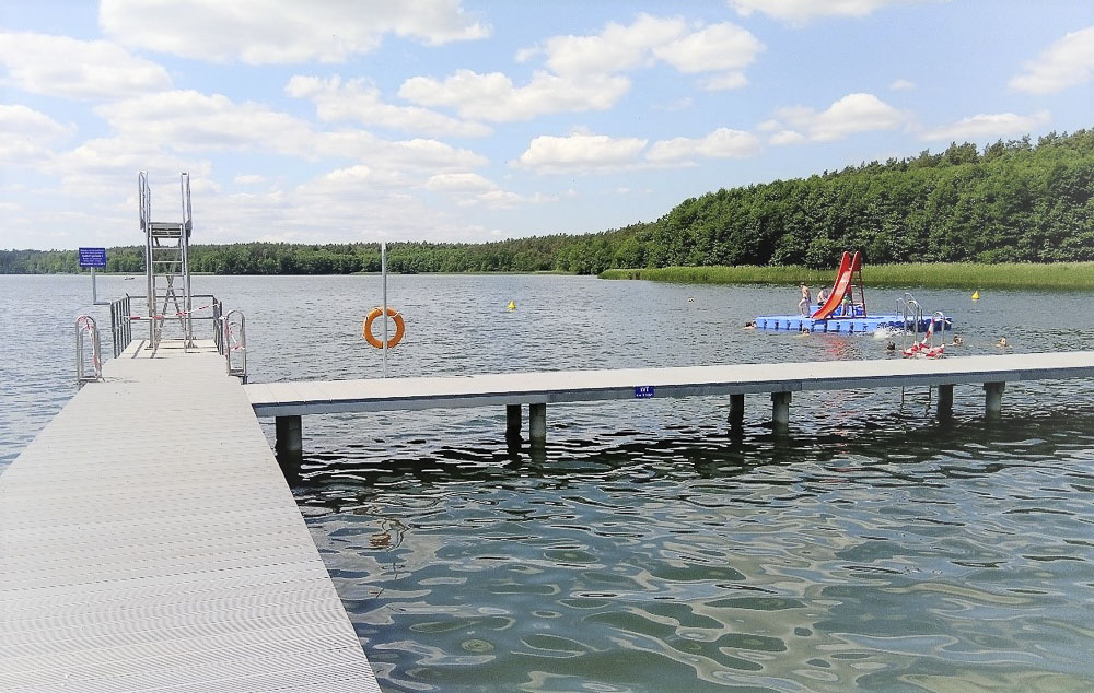 Ein weiter Blick über eine graue Steganlage mit einem Sprungturm auf das ruhige Wasser des Wolletzsees im Strandbad Wolletzsee. Rechts im See schwimmt eine blaue Plattform mit einer roten Wasserrutsche, umgeben von Badenden. Im Hintergrund erstreckt sich das grüne, bewaldete Ufer unter einem hellen Himmel mit vereinzelten Wolken.
