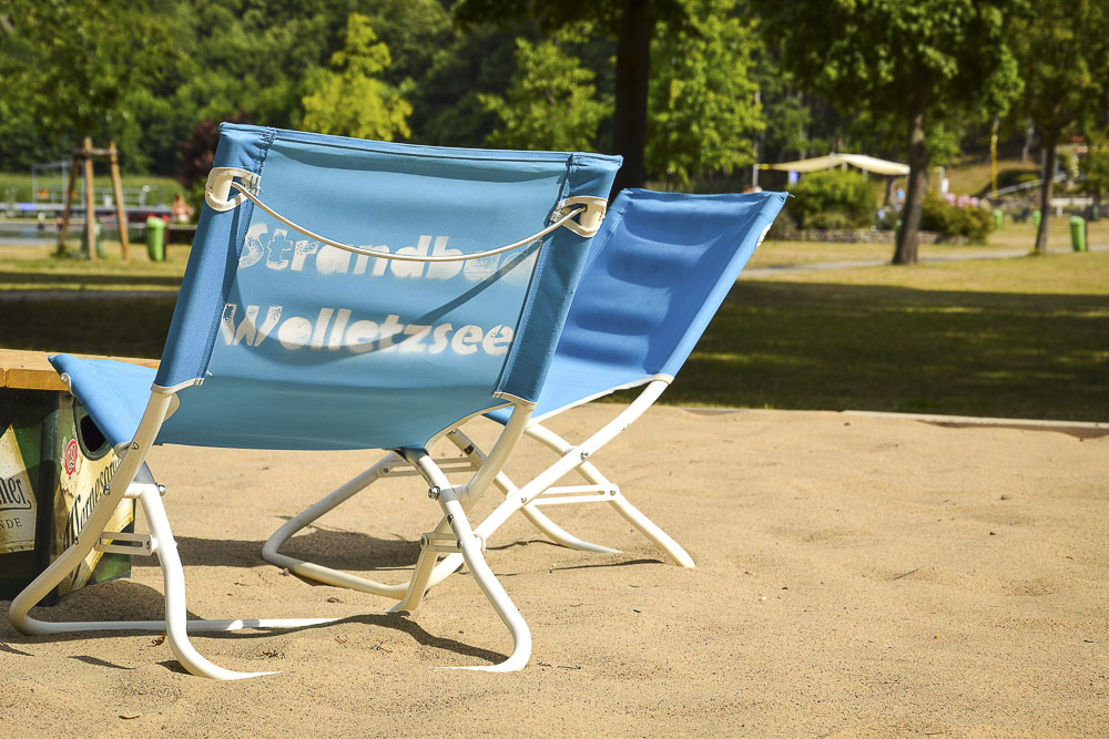 Zwei blaue Liegestühle mit weißem Metallgestell stehen auf einem sandigen Untergrund direkt im Strandbad Wolletzsee. Auf der Rücklehne des vorderen Liegestuhls ist der weiße Schriftzug „Strandbad Wolletzsee“ aufgedruckt. Im Hintergrund erstrecken sich eine begrünte Parkfläche mit Bäumen und eine sonnige Wiese.