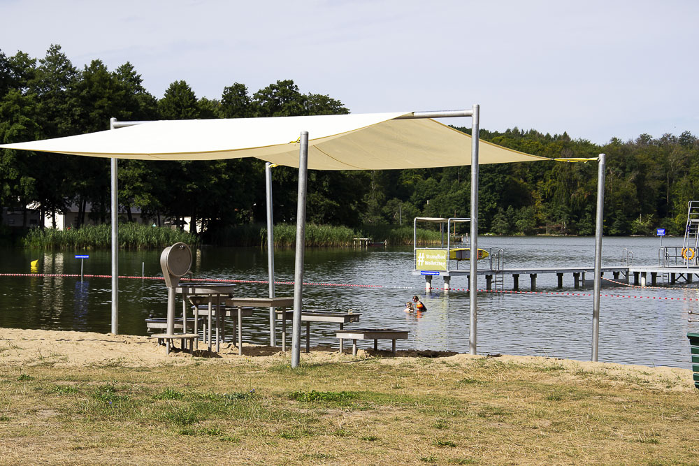 Ein von einem großen hellen Sonnensegel beschatteter Bereich mit Spieltischen steht direkt am Sandstrand des Strandbads Wolletzsee. Im Hintergrund sind das Wasser des Sees, Steganlagen sowie das bewaldete Ufer erkennbar.