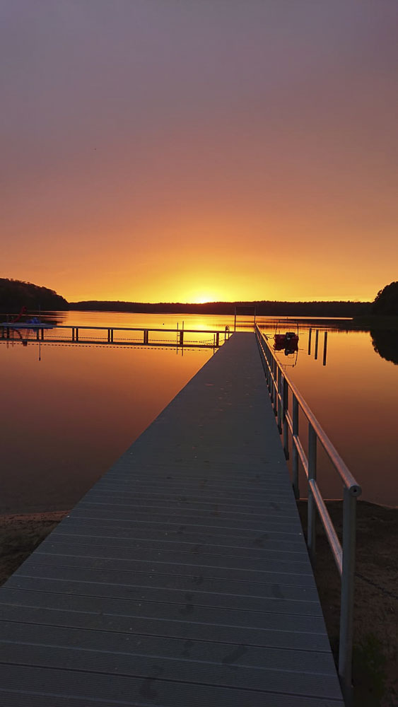 Ein malerischer Sonnenuntergang in kräftigen Orangetönen über dem Wolletzsee im Strandbad Wolletzsee. Im Vordergrund führt eine lange, mit einem Geländer versehene Steganlage geradewegs in das spiegelglatte Wasser, in dem sich der Abendhimmel reflektiert.