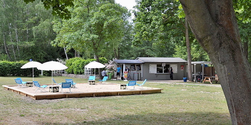 Beachbar im Strandbad Wolletzsee mit kleinen Tischen, blauen Liegestühlen und weißen Sonnenschirmen auf einer Sandfläche; im Hintergrund stehen Bäume und das Gebäude der Bar.