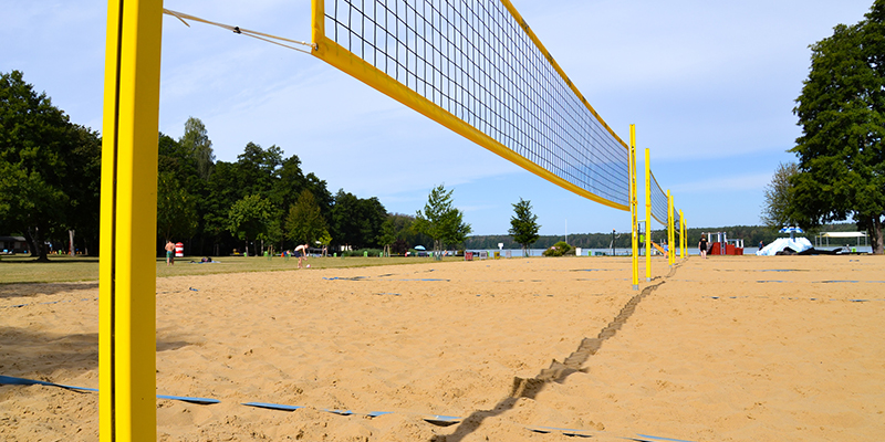 Beachvolleyballfeld mit gelbem Netz auf einer Sandfläche, im Hintergrund eine Liegewiese, Bäume und ein See.