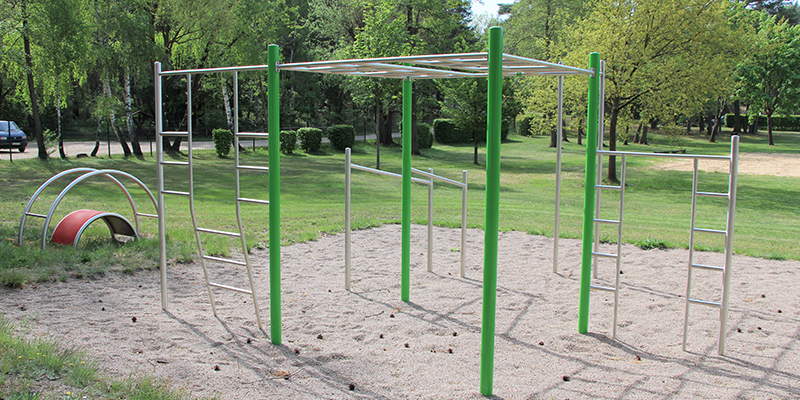 Outdoor-Fitnessanlage mit grünen Pfosten und silbernen Metallstangen auf einer Sandfläche, umgeben von Bäumen und Rasen