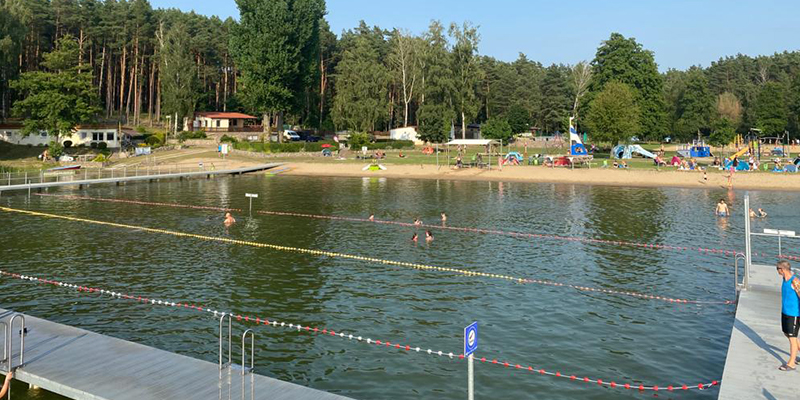Abgetrennter Schwimmbereich in einem See mit umlaufender Steganlage, durch Bojenketten markierten Wasserzonen und einem belebten Sandstrand im Hintergrund