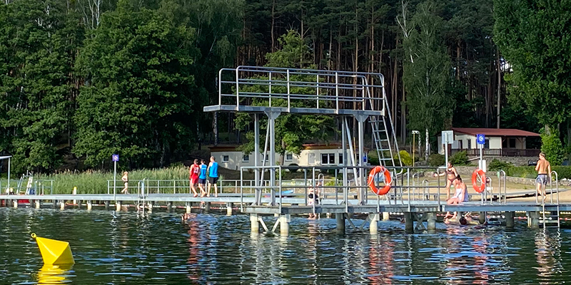 Ein metallener Sprungturm auf einem Steg in einem See, auf dem sich mehrere Badegäste aufhalten, umgeben von einem bewaldeten Ufer im Hintergrund.