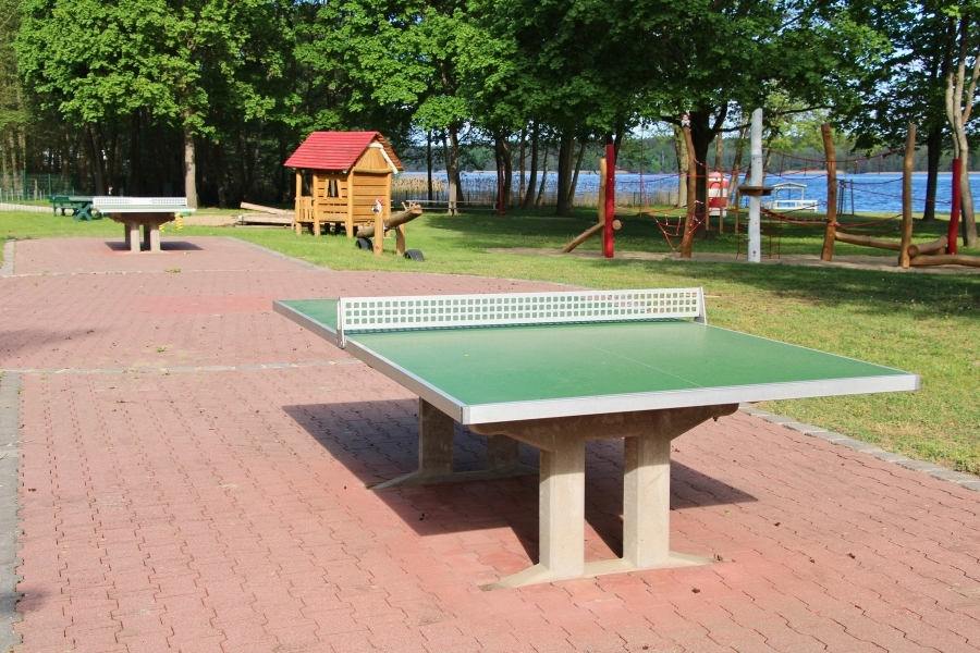Eine grüne Outdoor-Tischtennisplatte auf einer rötlich gepflasterten Fläche, im Hintergrund eine weitere Platte, hölzerne Spielgeräte und ein See.