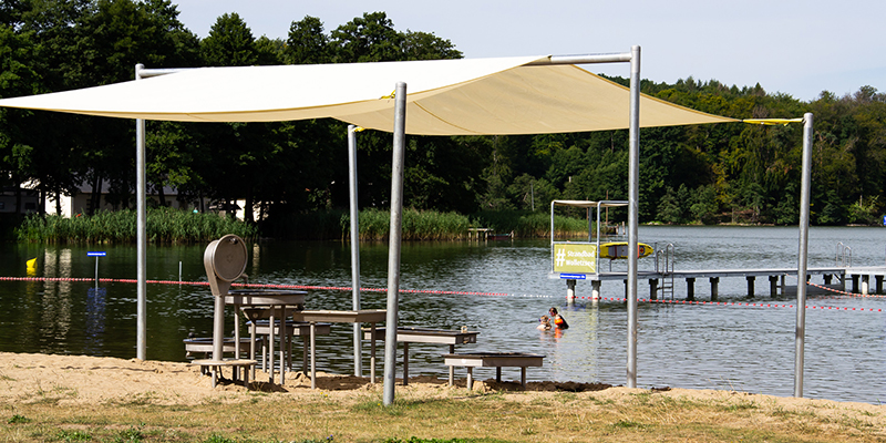 Ein von einem beigen Sonnensegel überdachter Wasserspielplatz am Sandstrand eines Sees, im Hintergrund eine Steganlage und ein bewaldetes Ufer.